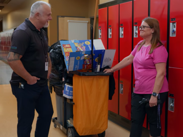 A male and female standing next to a custodian cart in a school hallway, having a conversation.